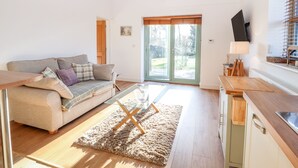 En moderne stue med sofa, sofabord i glas, et køkken med træbordplade og en skydedør af glas, der fører til en have.