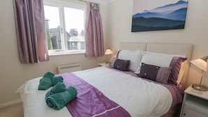 Un lit bien fait, recouvert d’une couvre-lit violette et blanche, avec deux oreillers et deux serviettes enroulées. Il y a une fenêtre avec des rideaux, un tableau accroché au mur et une table de chevet avec une lampe.