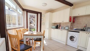 A compact kitchen with a washing machine, wooden chairs, a table, and a window overlooking a garden. - Ty Cerrig (Rhyl)