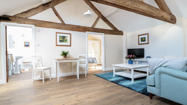 A spacious living room with wooden beams, a white sofa, a TV, and a dining area with a table and chairs. - Granary 2 (Hereford)