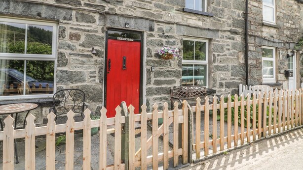 Un bâtiment en pierre avec une porte rouge portant l’inscription « PRESWYLEA », une clôture blanche en bois et une petite table avec un cadre en métal.