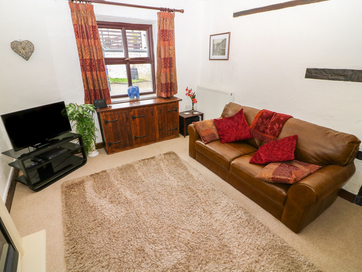 A living room with a brown leather sofa, a wooden cabinet, a television on a stand, and a window with curtains.