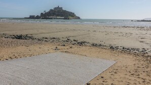 A beach with a castle-like structure in the distance, a sandy area with scattered rocks, and a paved section in the foreground. - Beachcomber (Marazion)