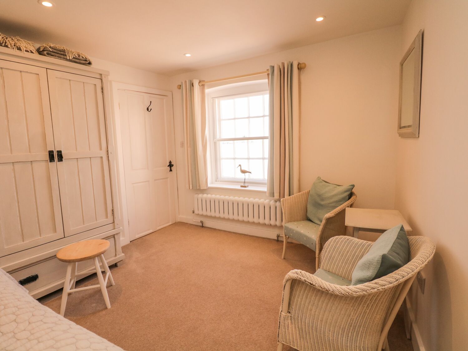 A bedroom with a large wardrobe, a small table, two wicker chairs, a radiator, and a window with curtains.