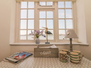 A window with a view of a body of water, a vase with flowers, a wooden box labeled "Apple," and two mugs on a table. - Searles (Exmouth)