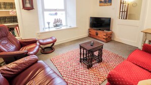 A living room with a leather sofa, a red patterned rug, a wooden coffee table, a television on a wooden cabinet, a bookshelf, and a window with a ship model. - Sea Spray (Eyemouth)