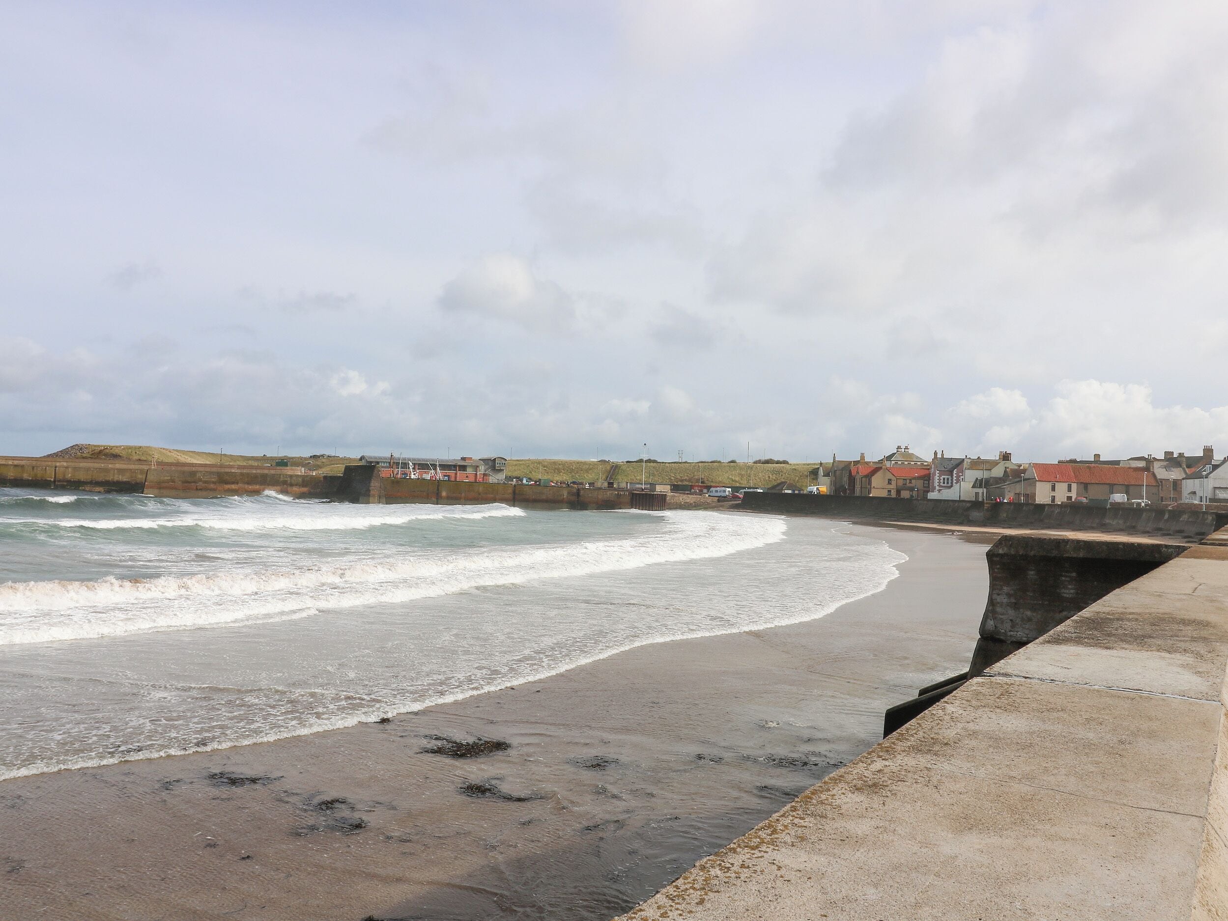 A coastal scene with a concrete sea wall, waves, and buildings in the background.