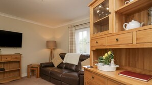 A cozy living room with a brown leather sofa, a wooden cabinet with glass doors, a flat-screen TV, a floor lamp, and a small side table. - Betty's Mews (Ambleside)