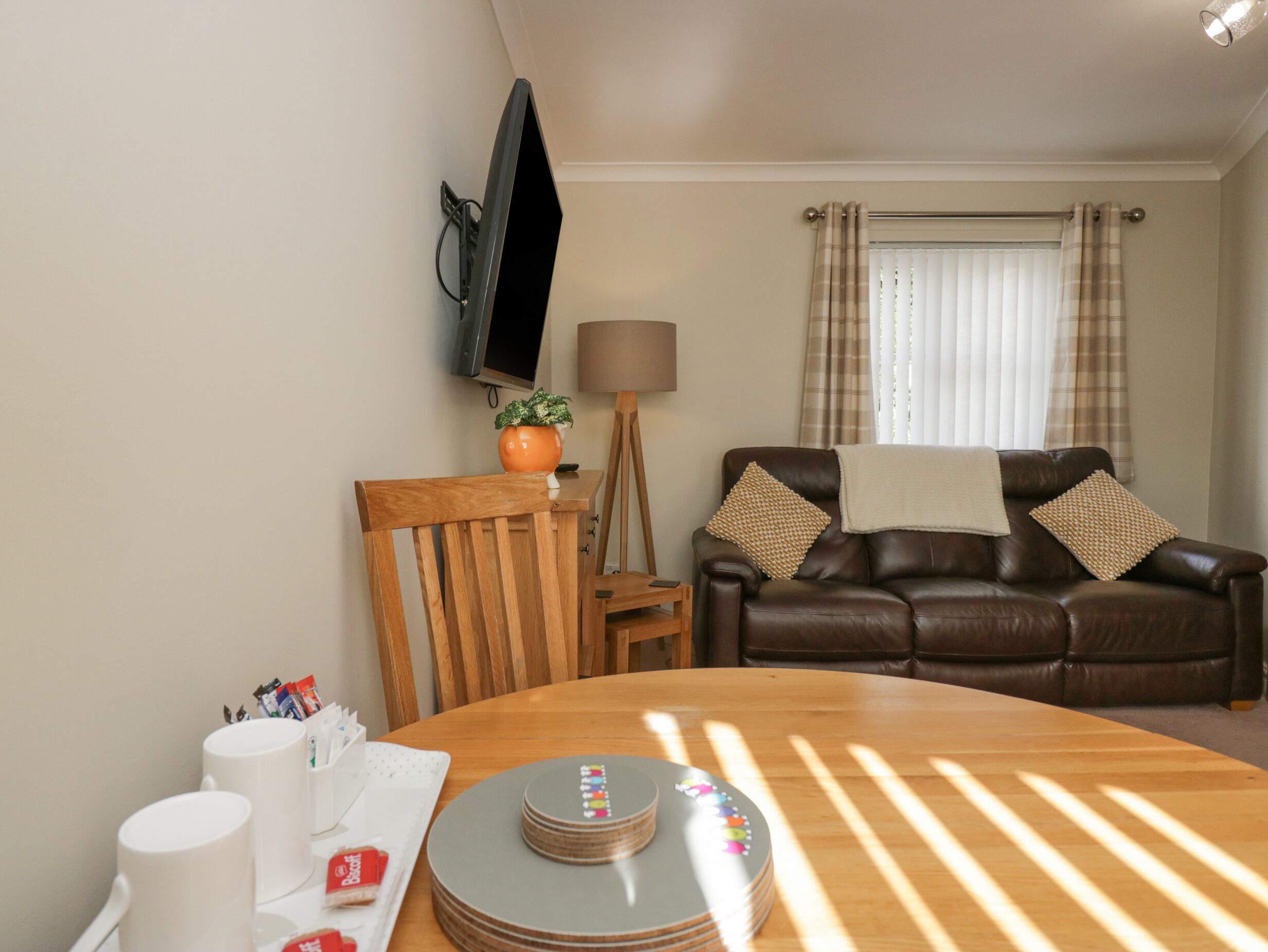 A living room with a wooden dining table, a brown leather sofa, a TV mounted on the wall, and a window with curtains.