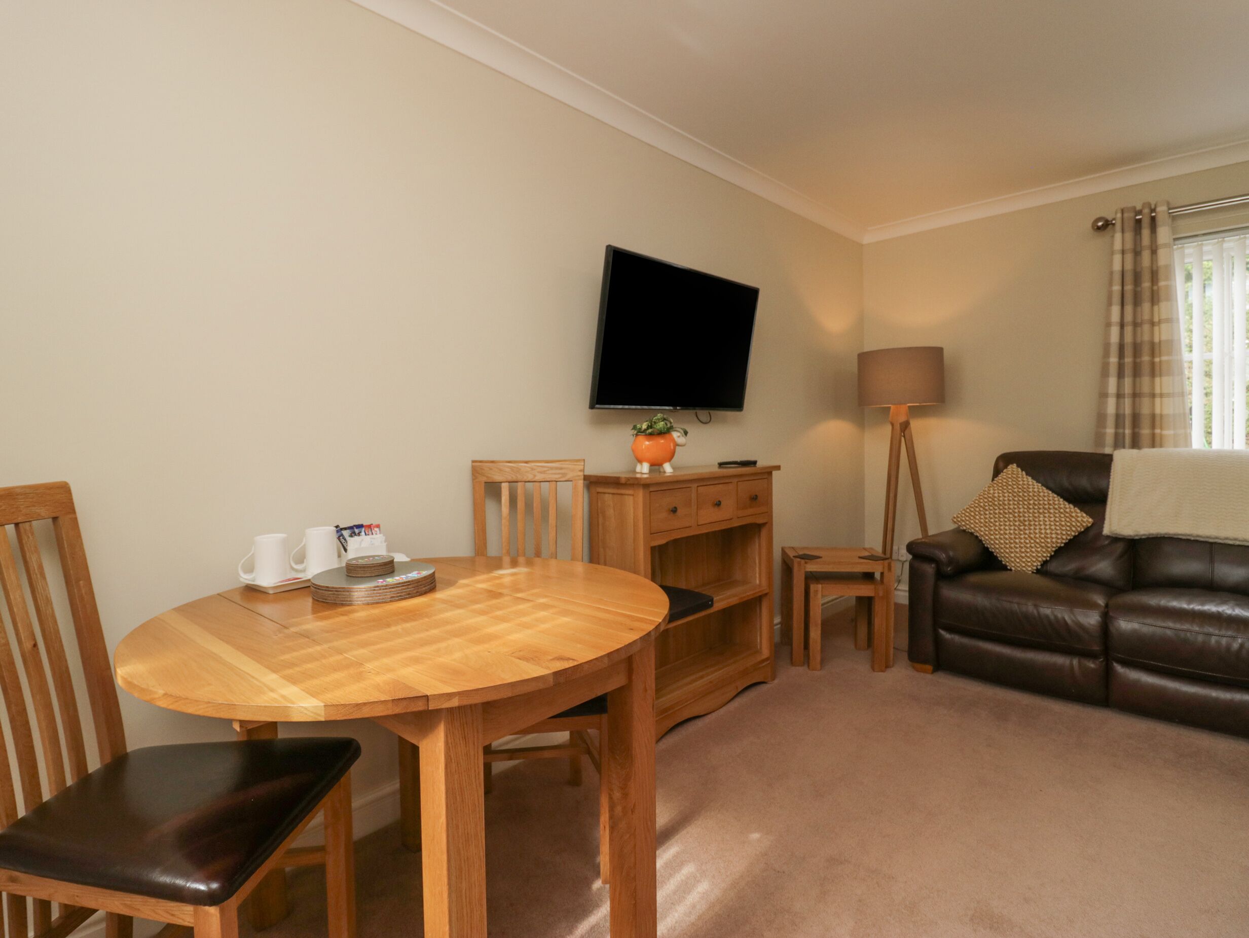 A cozy living room with a wooden dining table, chairs, a leather sofa, a TV mounted on the wall, and a floor lamp.