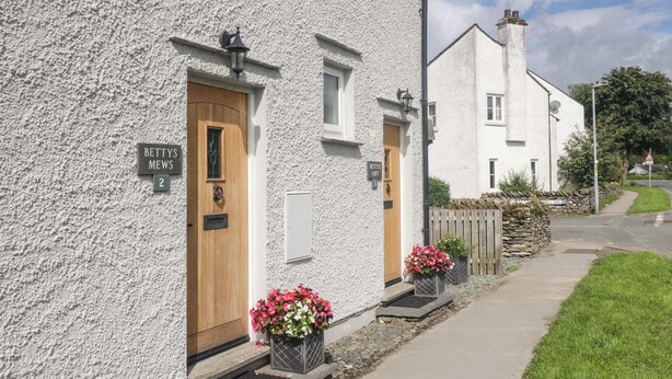 A white building with two wooden doors, each marked with a number. There are flower boxes with pink and white flowers on the steps. - Betty's Mews (Ambleside)