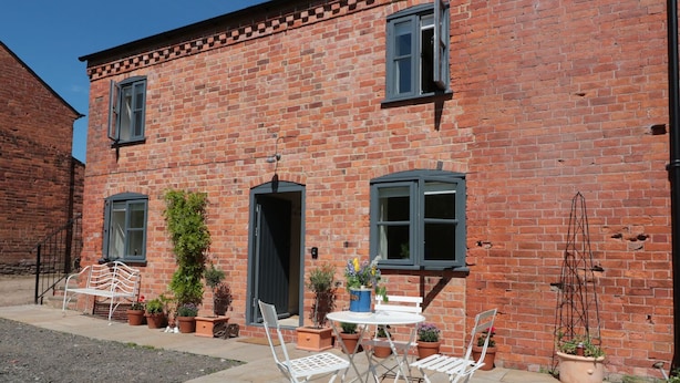 A brick building with a patio area featuring a table and chairs, potted plants, and a bench. - Granary 1 (Hereford)