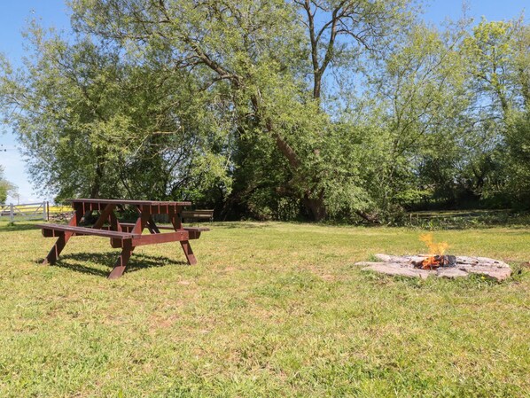 Una mesa de picnic y una fogata en un área con césped y árboles al fondo.