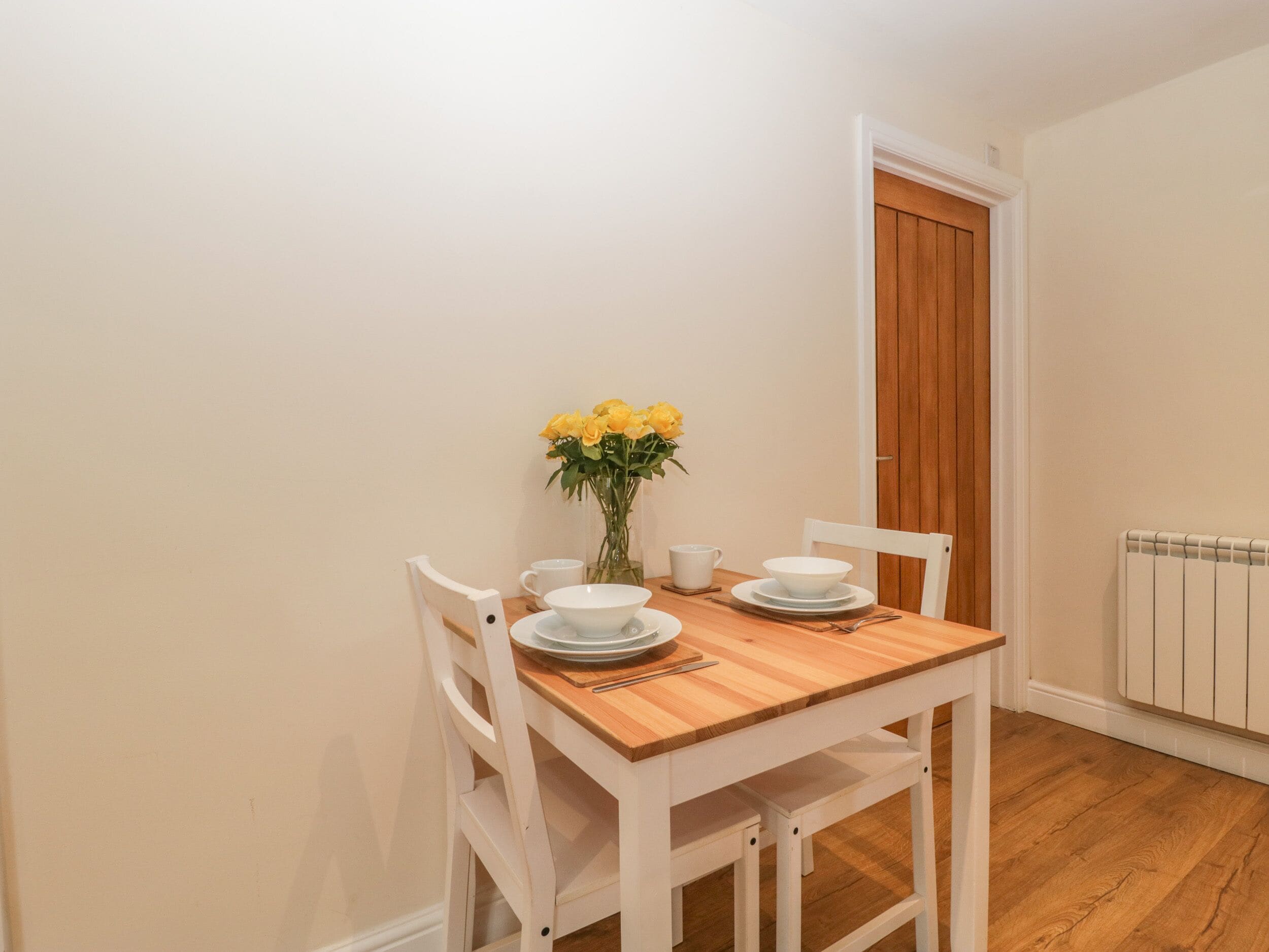 A dining area with a wooden table and white chairs, a vase of yellow flowers, and a white radiator in the background.