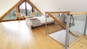 A bedroom with a sloped ceiling, a bed, two wicker chairs, and a glass railing. - Tyddyn Isa (Denbigh)