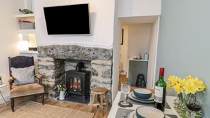 Ein gemütliches Wohnzimmer mit Kamin aus Stein, einem bequemen Sessel, einem Esstisch mit Geschirr und einer Weinflasche sowie einem an der Wand montierten Flachbildfernseher.