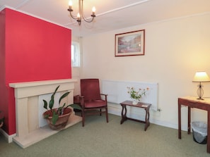A living room with a red wall, a fireplace, a red armchair, a small table with a vase of flowers, a lamp, and a radiator. - 1 Westcroft Cottage (Stonehouse)