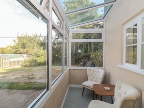 A room with a glass ceiling, a cushioned chair, a small table, and a view of a garden and trees. - 1 Westcroft Cottage (Stonehouse)