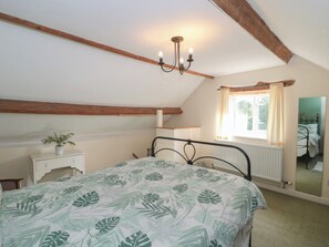 A bedroom with a sloped ceiling, a bed with a leaf-patterned bedspread, a bedside table with a plant, a window with curtains, and a mirror on the wall. - 1 Westcroft Cottage (Stonehouse)