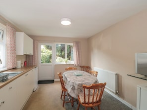 A compact kitchen with a dining area, a window with curtains, and a radiator. - 1 Westcroft Cottage (Stonehouse)