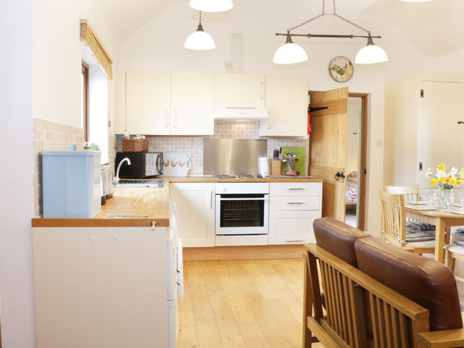 A compact kitchen with white cabinets, a blue refrigerator, a wooden dining table with chairs, and a clock on the wall.