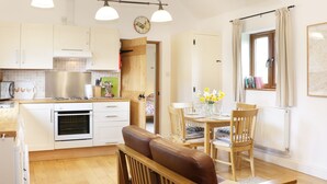 A cozy kitchen with wooden cabinets, a dining area with a table and chairs, and a window with curtains. - Acorn Cottage 1 (Shrewsbury)