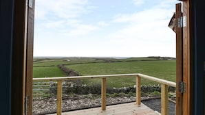 A wooden deck with a metal railing overlooking a green field and distant hills. - Cysgod y Bugail (Holyhead)
