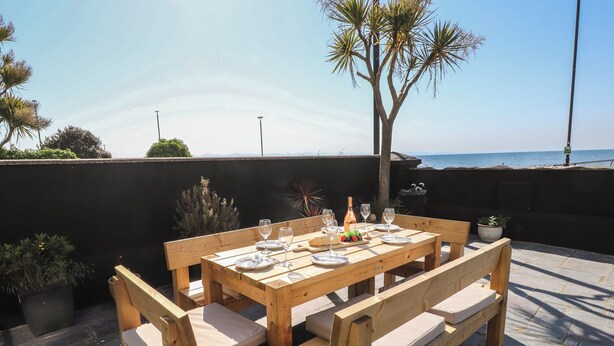An outdoor wooden table set with plates, glasses, and a bottle, surrounded by benches, with a view of the sea and palm trees. - Wavecrest (Pwllheli)