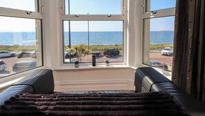 A room with a large bay window overlooking a beach, a sofa, and a small table with plants. - Wavecrest (Pwllheli)