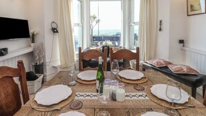 A dining room with a table set for four, large windows with curtains, a flat-screen TV mounted on the wall, and a view of the sea. - Wavecrest (Pwllheli)