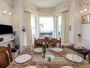 A dining room with a table set for four, large windows with curtains, a flat-screen TV mounted on the wall, and a view of the sea.