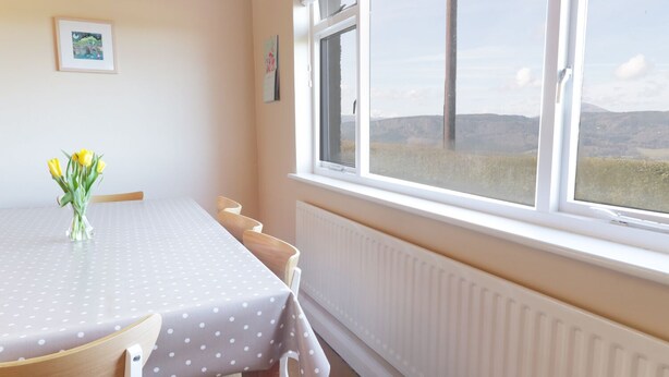 A dining area with a table covered by a polka dot tablecloth, a vase with yellow flowers, and a window with a view of hills. - Tai Candryll (Llanrwst)