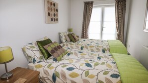 A bedroom with two beds, a bedside table, a lamp, and a window with curtains. - 24 Rhodewood House (Saundersfoot)