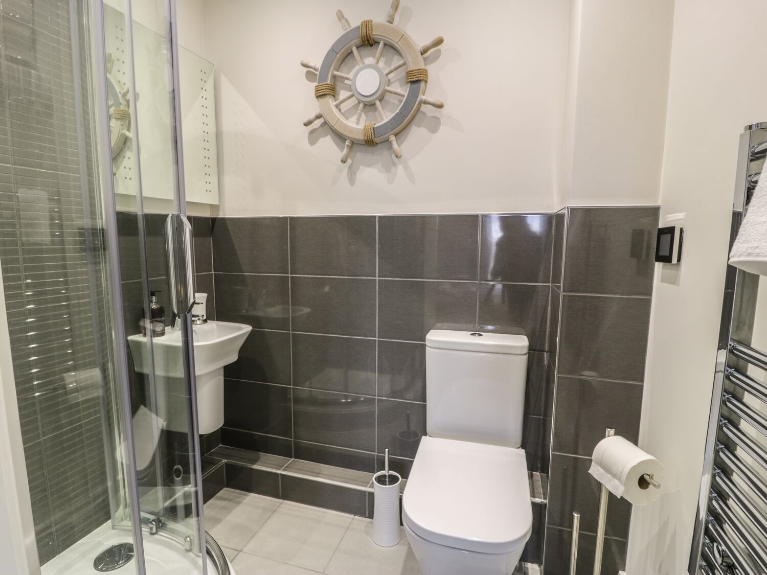 A modern bathroom with a glass shower enclosure, a white sink, a toilet, and a ship's wheel decoration on the wall.