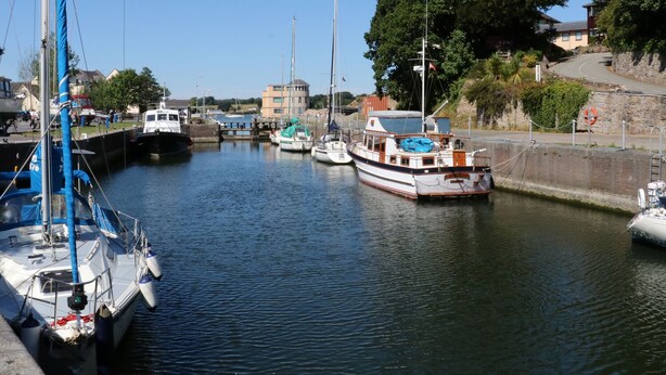 A marina with several boats docked, a stone wall, and a building in the background. - Glan Y Don (Y Felinheli)