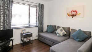 A living room with a grey sofa, a small table with books, a TV, and a window with patterned curtains. - Sea Breeze (Burnham-on-Sea)