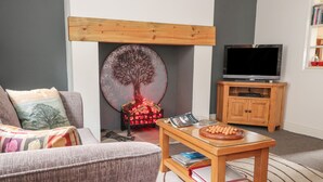 A living room with a sofa, a coffee table with books, a fireplace with a tree decoration, and a TV stand. - Heather Corner (Keighley)