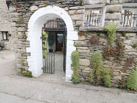 Un edificio de piedra con un arco de entrada blanco, una verja metálica y un muro de piedra cubierto de vegetación.