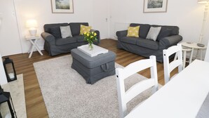 A modern living room with two grey sofas, a white coffee table, and a white dining table. There are framed pictures on the walls and a lamp on a side table. - Rose Cottage (Alnwick)