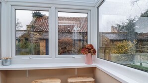 A bay window with two wooden stools, a pink vase with flowers, and a view of a stone building with a tiled roof. - Rose Cottage (Alnwick)