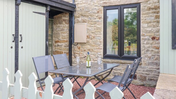 A stone building with a glass table and chairs, a white picket fence, and a window with a view of greenery.