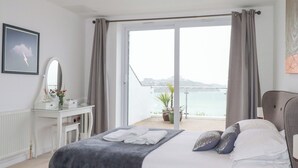 A bedroom with a bed, a bedside table, a mirror, and a view of the sea. - Buena Vista (Newquay)