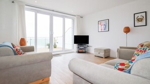 A spacious living room with a large sliding glass door leading to a balcony, a flat-screen TV, and a few pieces of furniture. - Buena Vista (Newquay)