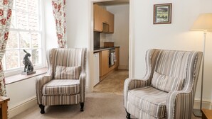 A room with two striped armchairs, a window with floral curtains, a statue, a framed picture, and a view into a kitchen area. - Spire View (Pickering)