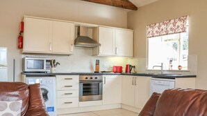 A compact kitchen with white cabinets, a microwave, a toaster, and a dishwasher. There is a window with floral curtains and a brown leather sofa in the foreground. - The Old Dairy (Worthing)