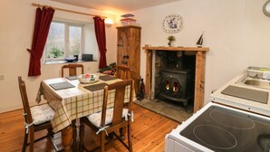 A cozy room with a fireplace, a dining table with chairs, a kitchen area with a stove, and a window with curtains. - Creag Mhor Cottage (Kyle)