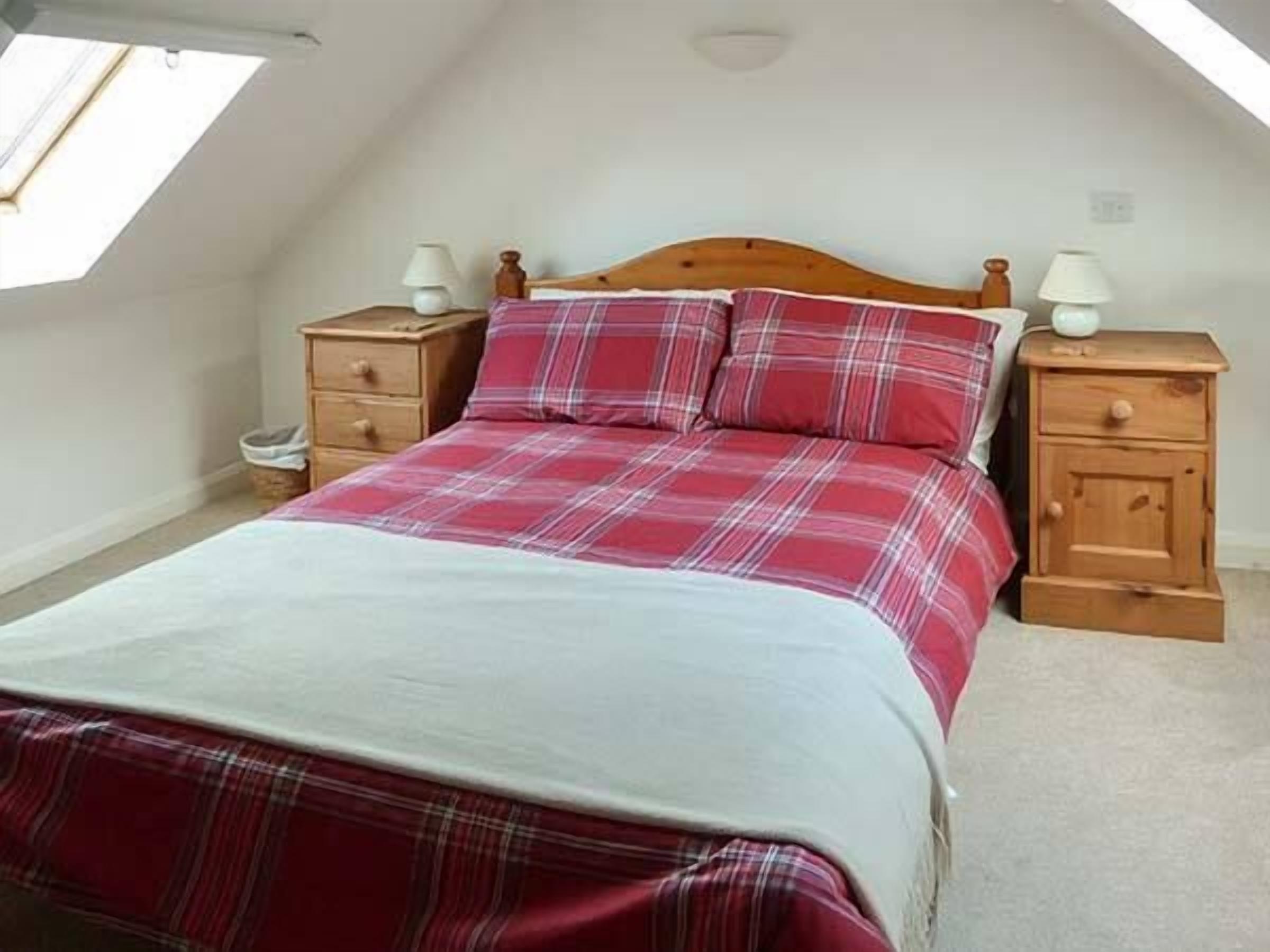 A bedroom with a sloped ceiling, a wooden bed frame, a red plaid bedspread, two bedside tables with lamps, and a skylight.