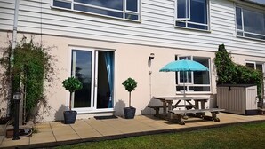 A two-story house with a patio area featuring a table and chairs, a blue umbrella, and potted plants. - Driftwood (Bude)
