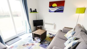 A living room with a grey sofa, a small wooden coffee table with magazines, a flat-screen TV, a colorful rug, and a painting on the wall. - Driftwood (Bude)