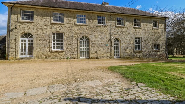 A stone building with multiple windows and a central door, surrounded by a gravel driveway and a grassy area.